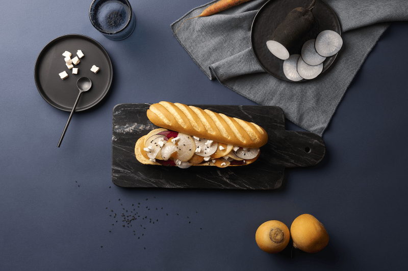 Photo d'une baguette viennoise sur une planche en marbre noir, garnis de tranches de radis noir et de carottes.