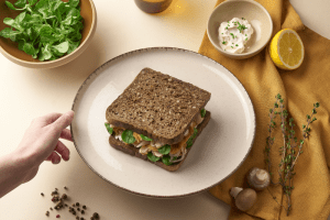 Un sandwich réalisé avec le pain de mie Seigle & Graines. Le tout est dans une assiette autour de laquelle il y a un demi-citron de la roquette et des champignons.