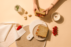 Photo de deux sandwichs garnis, un des deux est tenu par des mains et l'autre est dans une assiette au centre de la photo. Autour il y a des tomates, des poivrons et de l'ail.