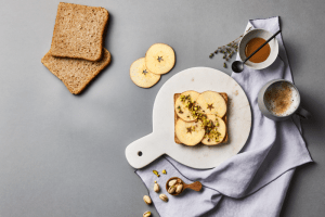 Une photo d'une tranche de pain de mie sur une planche en marbre. 4 tranches de pommes sont posées sur le pain de mie. Autour il y a des pistaches concassées, un café au lait ainsi que des branches de lavande.